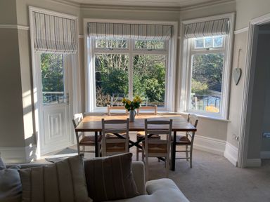 A dining area with a wooden table and six chairs by three windows with striped blinds and a door leading outside at The Anchorage in Bournemouth