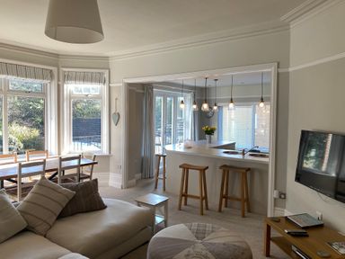 A living area with sofa and cushions dining table with chairs and kitchen counter with stools and hanging lights at The Anchorage in Bournemouth