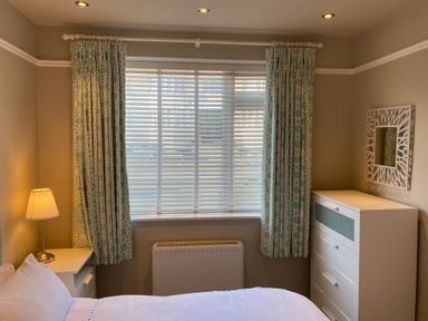 A bedroom with a bed a nightstand with a lamp a window with blinds and curtains a white chest of drawers and a decorative mirror on the wall at The Anchorage in Bournemouth