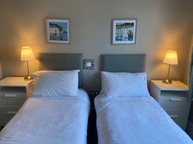 A bedroom with two single beds each with a white pillow and sheet bedside tables with lamps and framed artwork on the wall at The Anchorage in Bournemouth