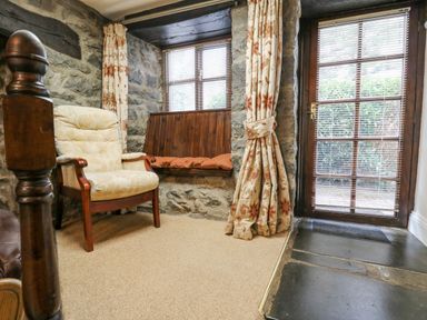 A corner of a room with a beige padded chair wooden bench under windows and a glass door with floral curtains at The Searle in Nantmel near Rhayader