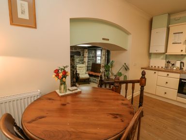 A wooden dining table with a vase of flowers next to a kitchen with white cabinets and an opening to a living area with stone walls and a chair at The Searle in Nantmel near Rhayader