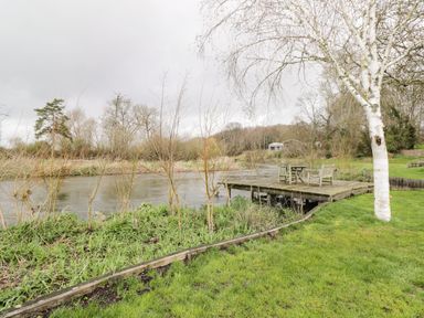 A wooden deck with chairs and a table beside a river with trees and grass at Avon Turn View in Alderbury