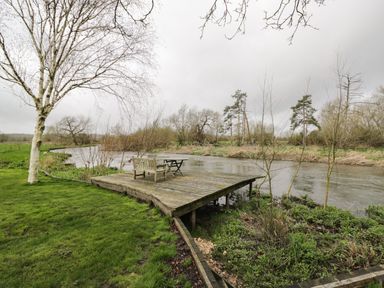 A wooden deck with a bench and table by a river with trees and grass at Avon Turn View in Alderbury