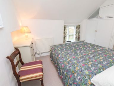 A bedroom with a bed covered by a floral quilt a wooden chair with a striped cushion a white dresser with a lamp and a window with floral curtains at Riseholme in Ebberston