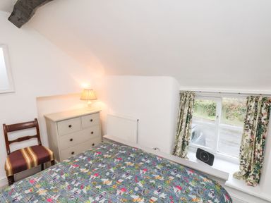 A bedroom with floral bedspread a wooden chair a chest of drawers with a lamp and a window with floral curtains at Riseholme in Ebberston