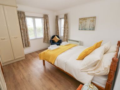 A bedroom with a double bed with yellow and white bedding a blue armchair with a yellow cushion patterned curtains wooden flooring and a wardrobe at Timaru in Lamphey