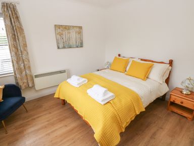 A bedroom with a wooden bed with yellow bedding and pillows a wooden nightstand with a lamp a blue chair and a window with patterned curtains at Timaru in Lamphey