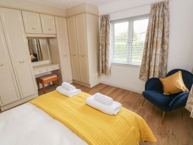 A bedroom with built in wardrobes a dressing table a bed with a yellow cover and white towels a blue chair and a window with patterned curtains at Timaru in Lamphey
