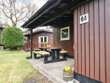 A covered outdoor seating area with a wooden picnic table and benches with cushions and yellow flowers at 64 Penlan Holiday Park in Cenarth