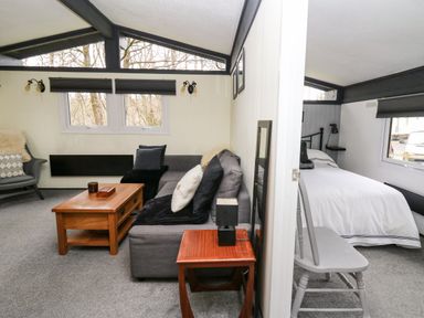 A living room with a gray sofa, wooden coffee table, and chair next to a bedroom with a single bed and chair at 64 Penlan Holiday Park in Cenarth