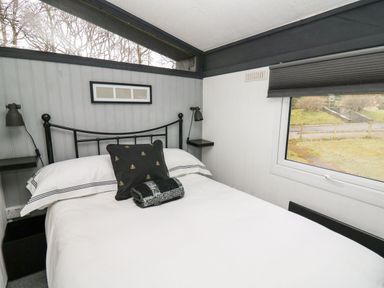 A bedroom with a black metal bed frame white bedding a black pillow a rolled blanket wall lamps and a window showing outdoor greenery at 64 Penlan Holiday Park Cenarth