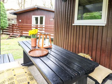 An outdoor wooden picnic table with cushions a flower pot and lantern near a wooden cabin at 64 Penlan Holiday Park Cenarth