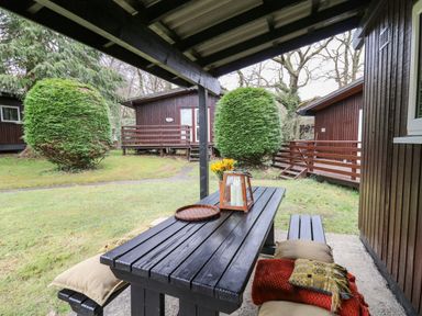 An outdoor covered seating area with a black wooden table and benches with cushions and a lantern surrounded by grass and wooden cabins at 64 Penlan Holiday Park in Cenarth