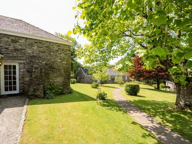 A garden with stone buildings trees and a paved pathway at Byre Trethin near Camelford