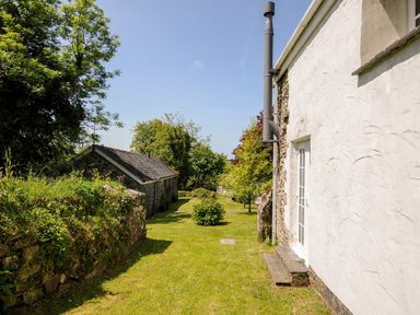 A garden path between a white house wall and a stone building with trees and grass at Byre Trethin near Camelford