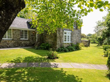 A stone house with white framed windows and a slate roof surrounded by a green lawn and trees at Byre Trethin near Camelford