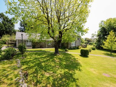 A garden with trees and a stone house with white windows at Byre Trethin near Camelford