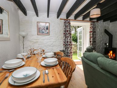 A dining area with a wooden table set with dishes and glasses next to a living area with green sofas and a wood stove at Byre Trethin near Camelford