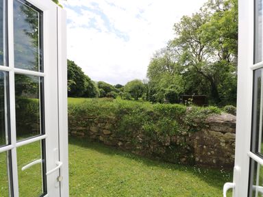 A view through open glass doors showing a garden with grass and a stone wall with trees beyond at Byre Trethin near Camelford
