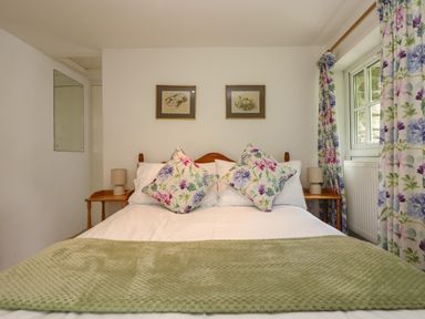 A bedroom with a bed with floral pillows and a green blanket beside two wooden bedside tables with lamps at Byre Trethin near Camelford