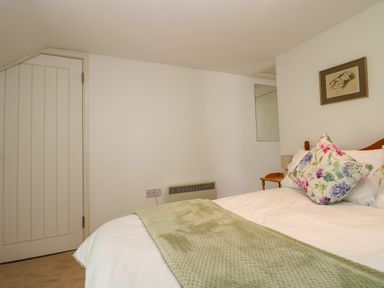 A bedroom with a bed covered in white sheets and a green blanket with floral cushions a door on the left and a chair in the corner at Byre in Trethin near Camelford