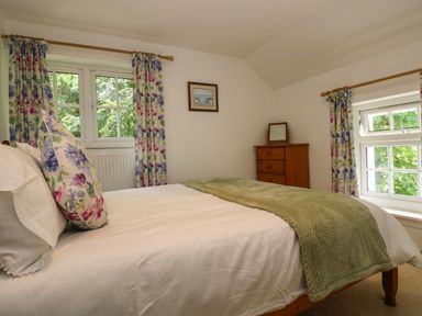 A bedroom with a bed with floral pillows and green blanket near two windows with floral curtains and a wooden chest of drawers at Byre Trethin near Camelford