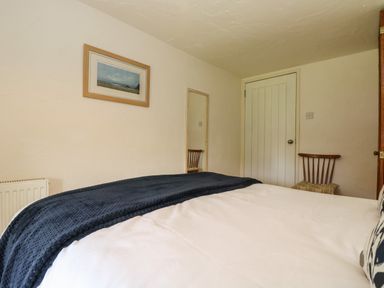 A bedroom with a bed covered by white and dark blue bedding a wooden chair with a cushion a wall mirror a framed picture and a closed door at Byre Trethin near Camelford