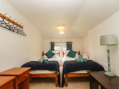 A bedroom with two single beds side by side each with pillows and towels a window with curtains a bedside table with a lamp and a wooden drawer unit with coat hangers on the wall at Byre in Trethin near Camelford