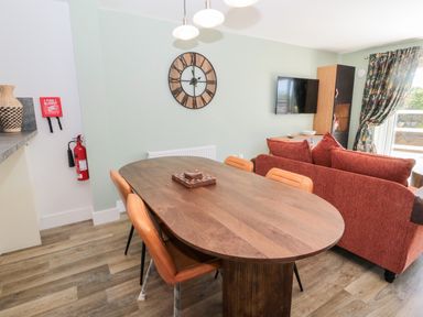 A dining area with a wooden table and four chairs next to a living room with a red sofa and a wall-mounted TV at Thrift Lodge in High Newton-by-the-Sea near Embleton