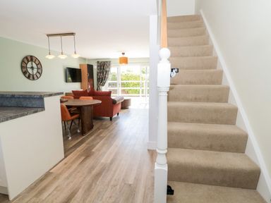 A living room and dining area with a round table and chairs a wall clock TV and sofa near carpeted stairs at Thrift Lodge High Newton-by-the-Sea near Embleton