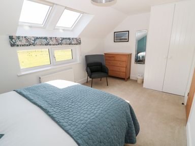 A bedroom with a bed covered in a blue quilt a dark grey chair a wooden chest of drawers a mirror a white wardrobe and windows at Thrift Lodge in High Newton-by-the-Sea near Embleton