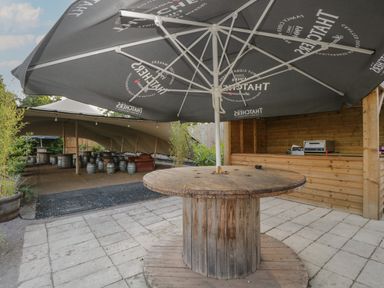 An outdoor seating area with a wooden spool table and umbrella near a wooden bar and covered seating area at Beech in Kemble