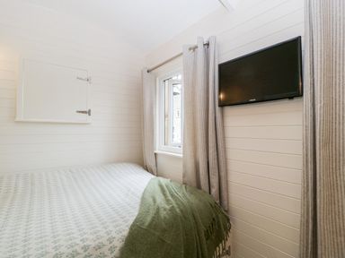 A bedroom with a bed covered with a patterned bedspread and green blanket a window with curtains and a wall mounted television at Hazel in Kemble