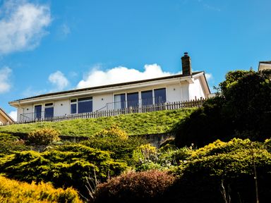 A white house with large glass windows on a grassy hill surrounded by bushes at Harbour Lights Port Isaac