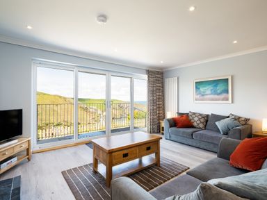 A living room with two grey sofas cushions a wooden coffee table carpet tv on a wooden stand and sliding glass doors with a view at Harbour Lights Port Isaac