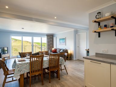 A dining area with a wooden table and chairs next to a living room with a sofa and sliding glass doors showing a hill outside at Harbour Lights Port Isaac
