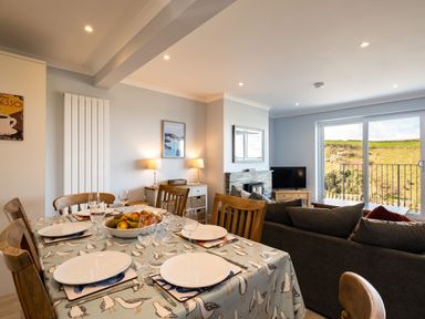 A dining area with a table set for six and a living area with a sofa a fireplace and a TV with a large window showing a hillside at Harbour Lights Port Isaac