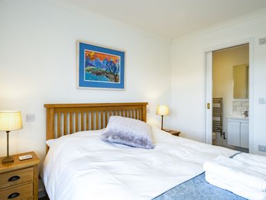 A bedroom with a wooden bed frame white bedding two bedside tables with lamps and a bathroom visible through a sliding door at Harbour Lights Port Isaac