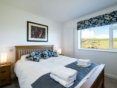 A bedroom with a wooden double bed white bedding folded towels bedside tables with lamps and a window showing a hill at Harbour Lights Port Isaac