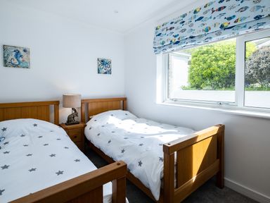 A bedroom with two single wooden beds white star patterned bedding a bedside table with a lamp and window with fish patterned blind at Harbour Lights Port Isaac