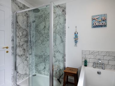 A bathroom with a glass shower enclosure, a bathtub, a wooden stool, and decorative items on the wall at Harbour Lights Port Isaac