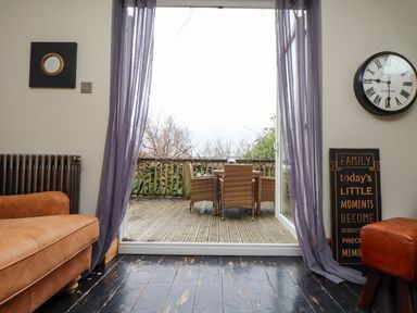 An indoor room with a brown leather sofa and matching footstool next to an open door leading to an outdoor wooden deck with a wicker table and chairs at Hillcroft in St Austell
