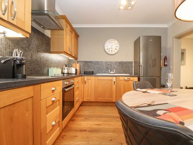 A kitchen with wooden cabinets a round dining table black chairs a clock on the wall and a refrigerator at Hillcroft in St Austell