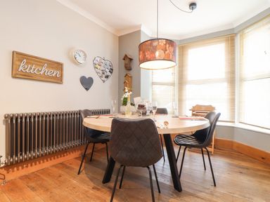 A dining area with a round wooden table surrounded by four black chairs under a pendant light with blinds on the windows at Hillcroft in St Austell