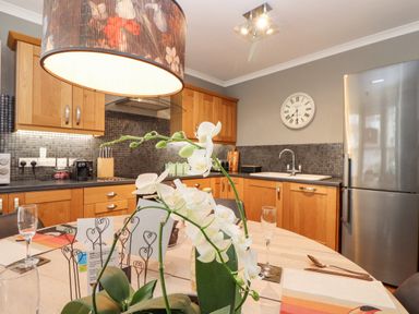 A kitchen with wooden cabinets a stainless steel refrigerator a wall clock and a dining table with white flowers at Hillcroft in St Austell