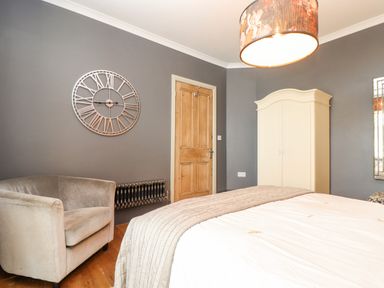 A bedroom with a bed with white bedding a beige armchair a wooden door a white wardrobe and a wall clock at Hillcroft in St Austell