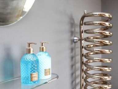 Blue soap and lotion bottles on a glass shelf with a round mirror and a metal spiral towel warmer in a bathroom at Hillcroft in St Austell
