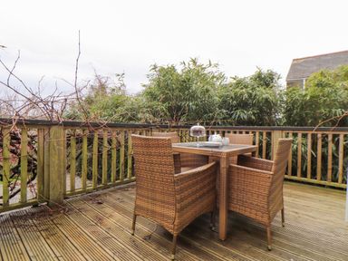 An outdoor wooden deck with a wicker table and four matching chairs surrounded by greenery at Hillcroft in St Austell