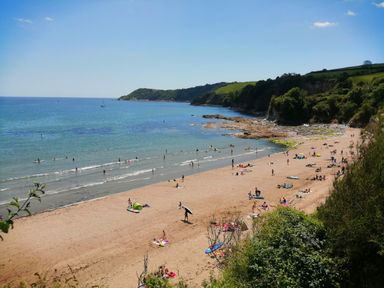 A beach with people sunbathing swimming and surfing near cliffs and greenery at Hillcroft in St Austell